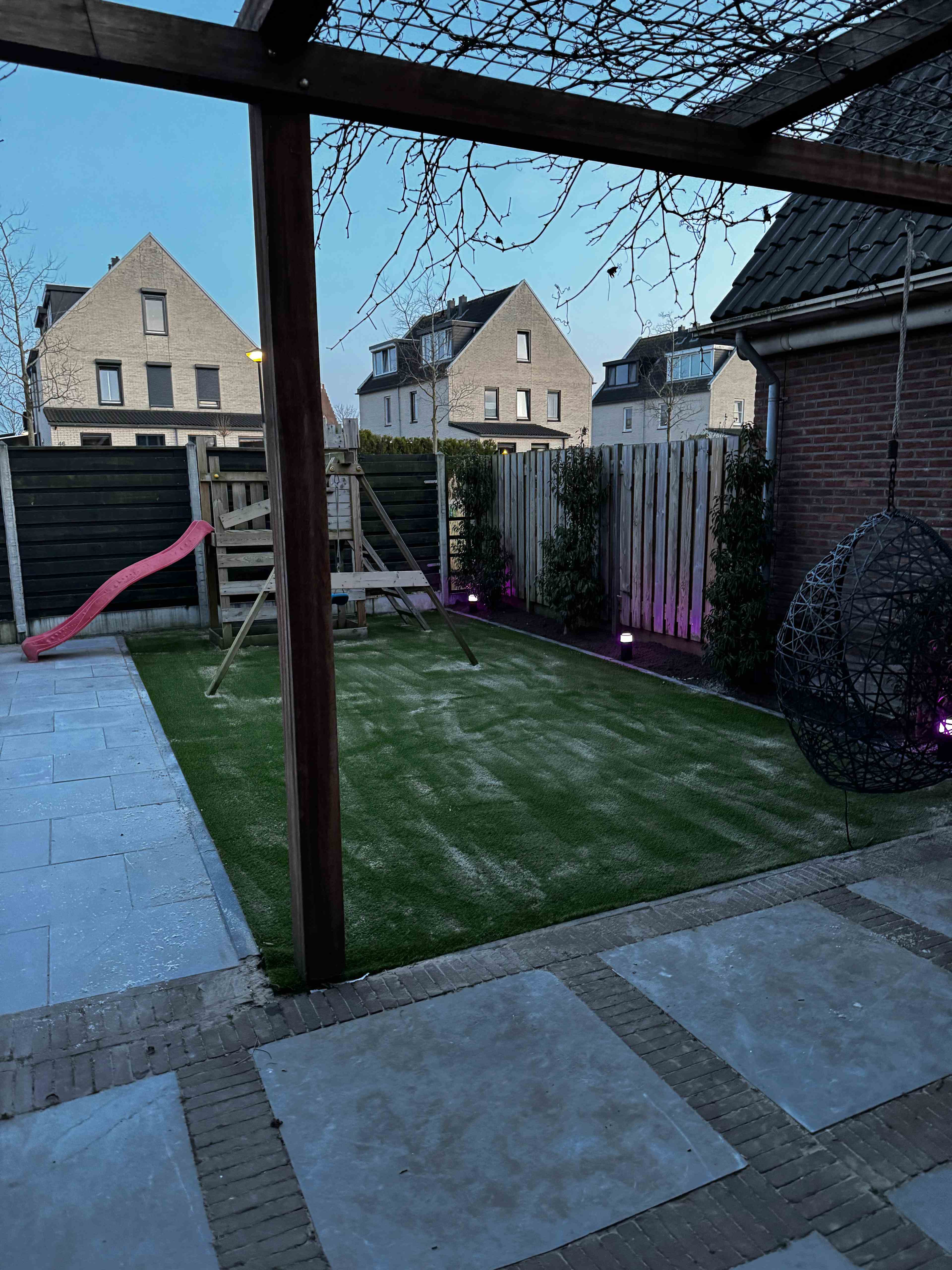 Aangelegde tuin met kunstgras, bestrating, pergola en hangstoel in de schemering