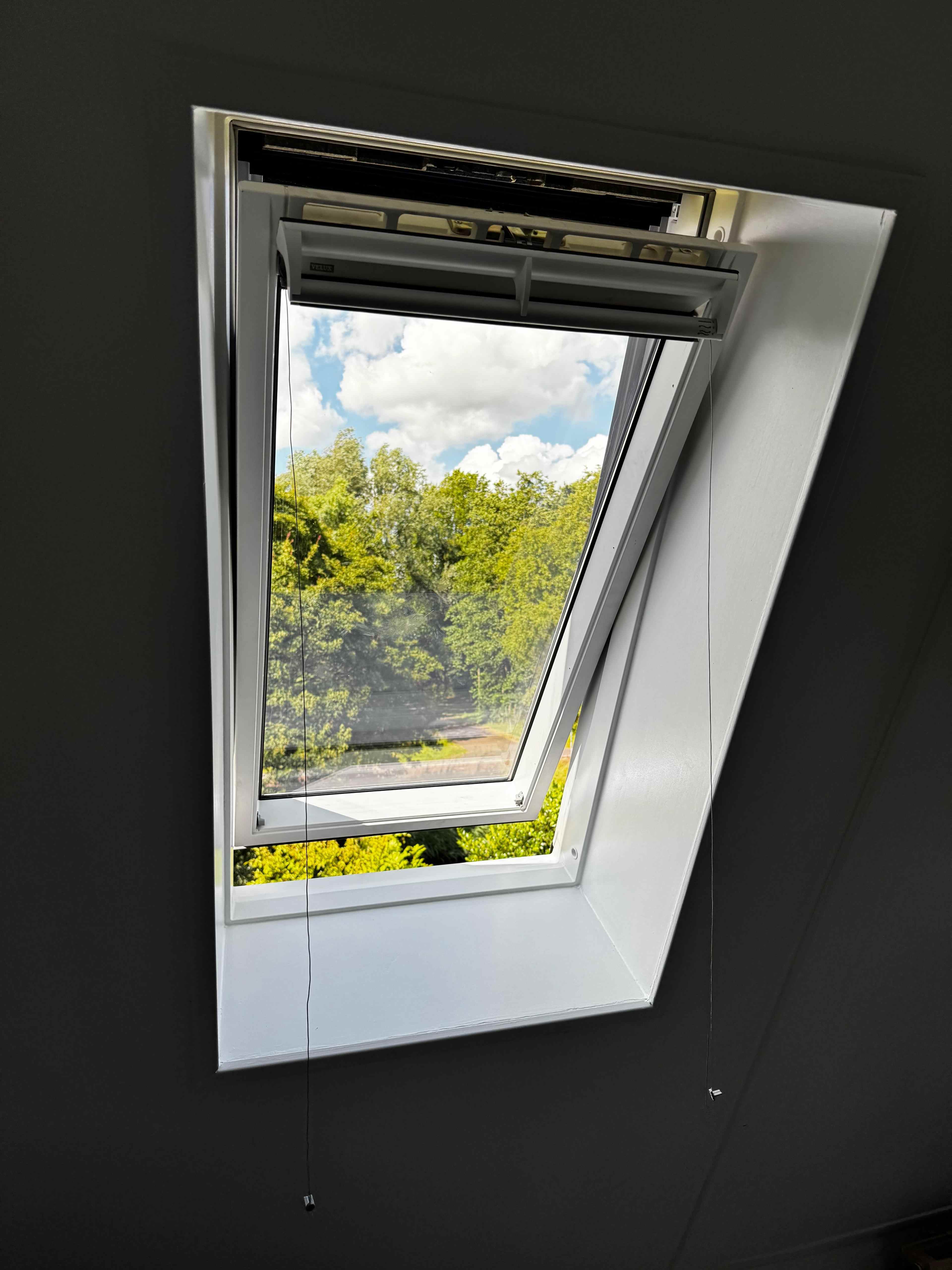 Wit dakraam geopend met uitzicht op groene bomen en blauwe lucht